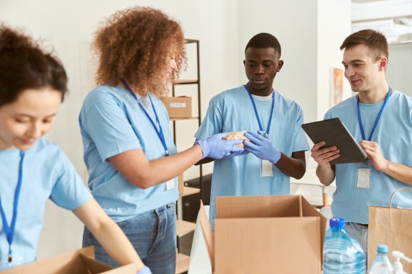 Diverse young volunteers in protective gloves sorting, packing foodstuff in cardboard boxes, working